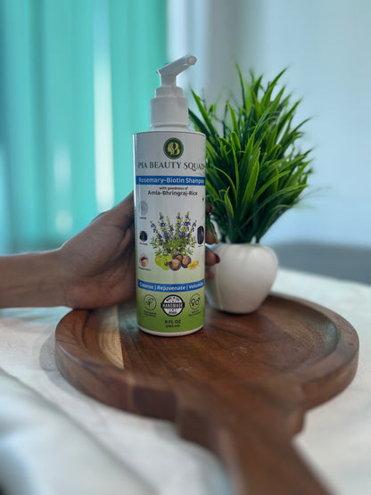 A bottle of handmade Rosemary Biotin Shampoo by PIA Beauty Squad on a wooden tray, with a white ball and green plant in the background.